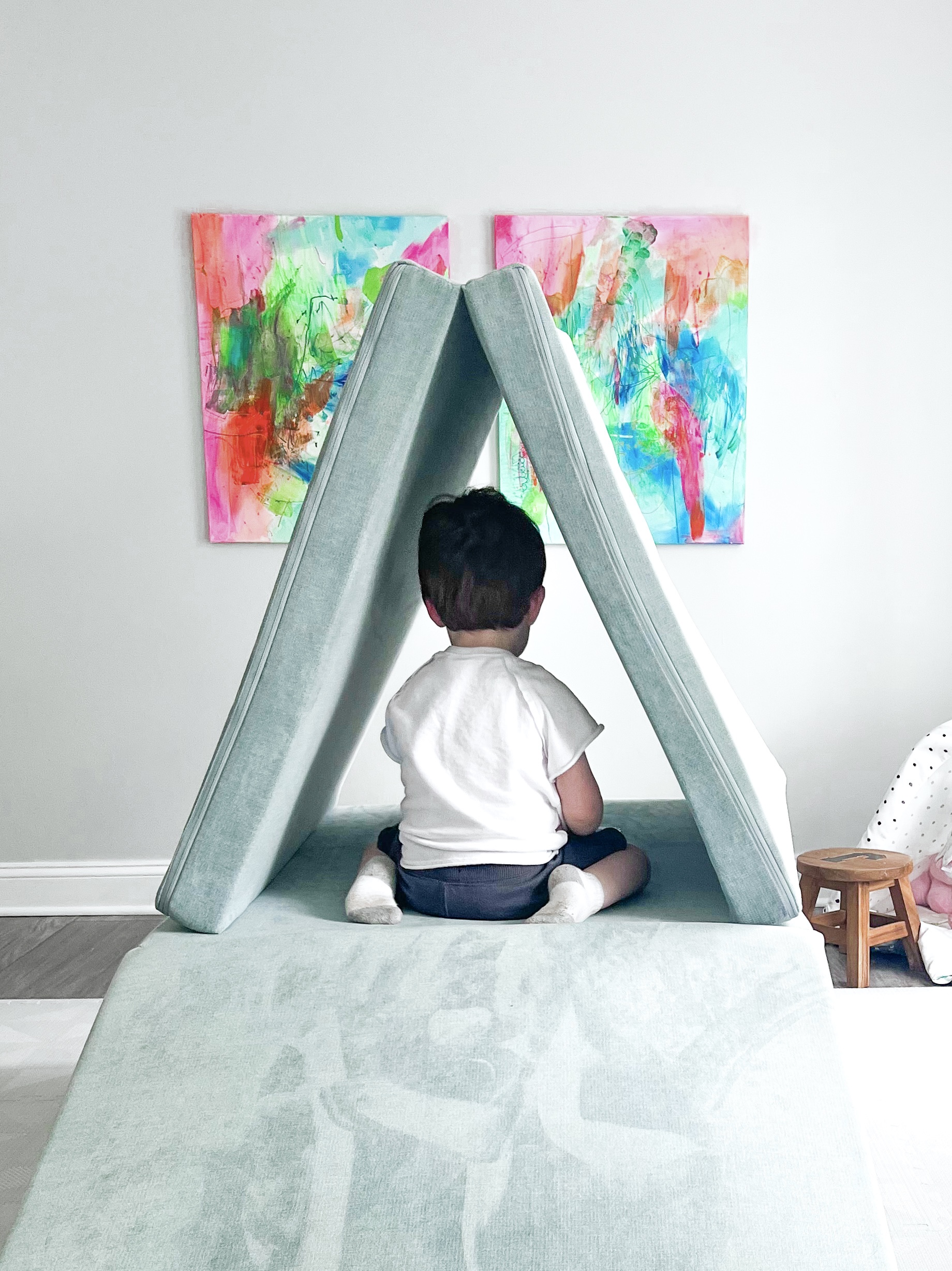Pale blue Nugget play couch in a teepee formation with small child inside. Abstract paintings on wall in background