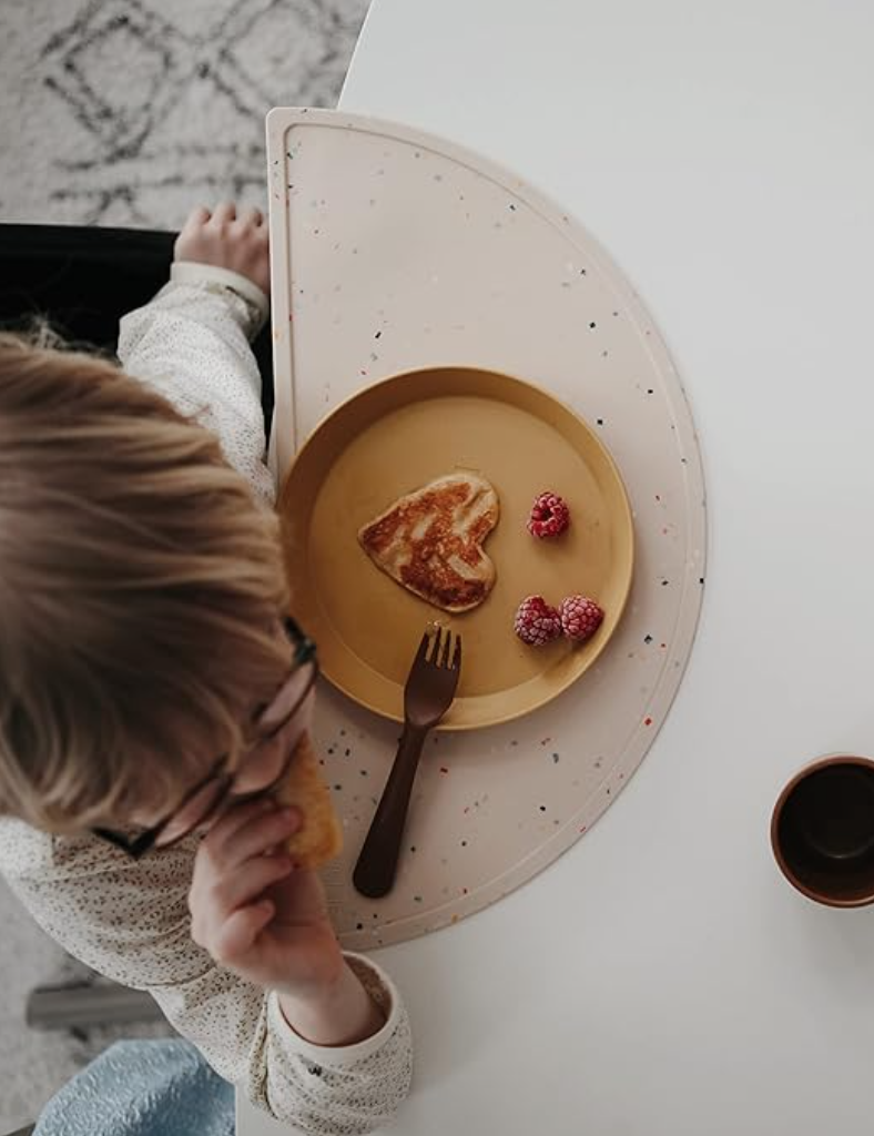Overhead shot of a little boy eating a heart shaped pancake on yellow plate with Mushi brand placemat