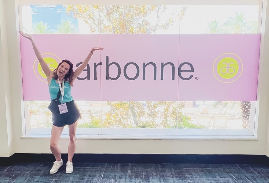 White woman with brown hair smiling with arms raised in front of an Arbonne sign on a large window.