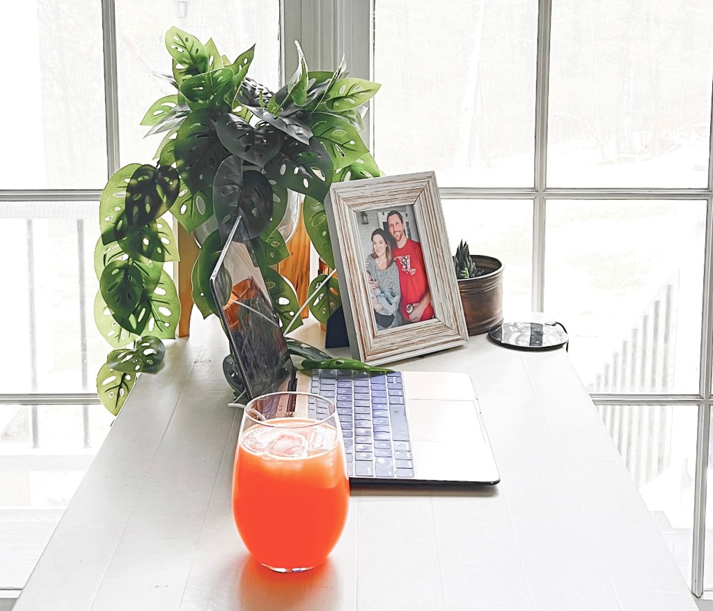 Orange Energy Fizz in stemless wine glass on table, in front of a laptop, family photo, plant, and bright window